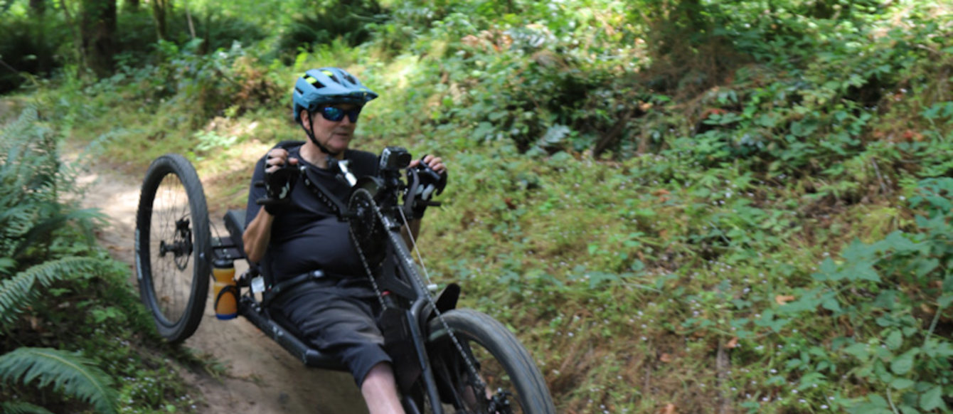 Max Woodbury rides an adaptive mountain bike down a dirt path in woods