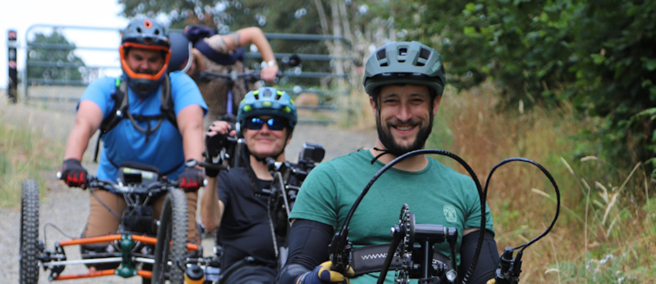 three adaptive mountain bikers, smiling, riding down a gravel path