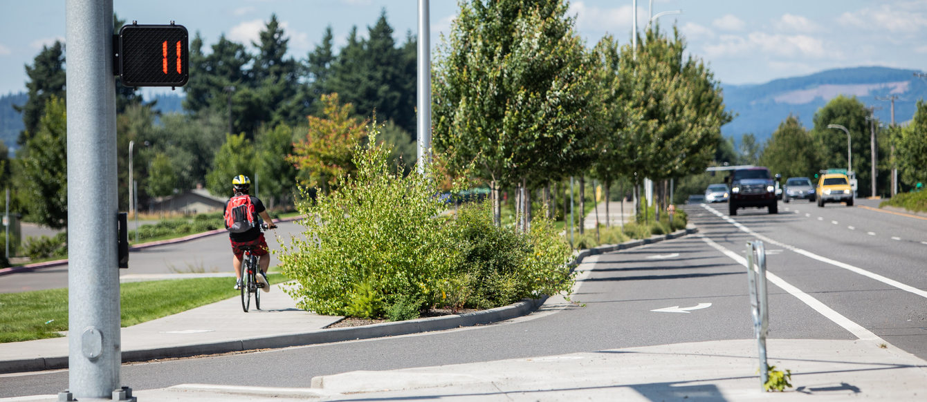 an intersection in Gresham, Oregon on Northeast Hogan Drive. There is a crosswalk with the walk countdown meter at 11. Cars are approaching on the right side. On the left a biker is riding on a biking and walk path. Trees line the street.