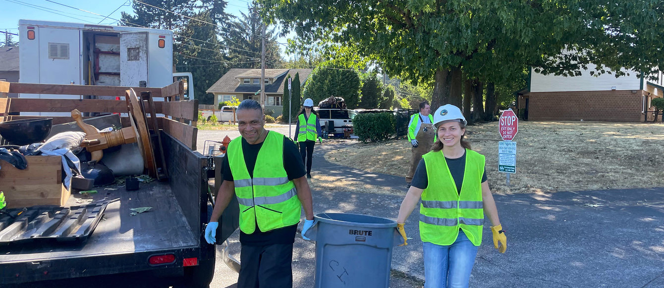 A man and woman carry a trash can to an open trailer bed. Both wear neon yellow high visibility vests.