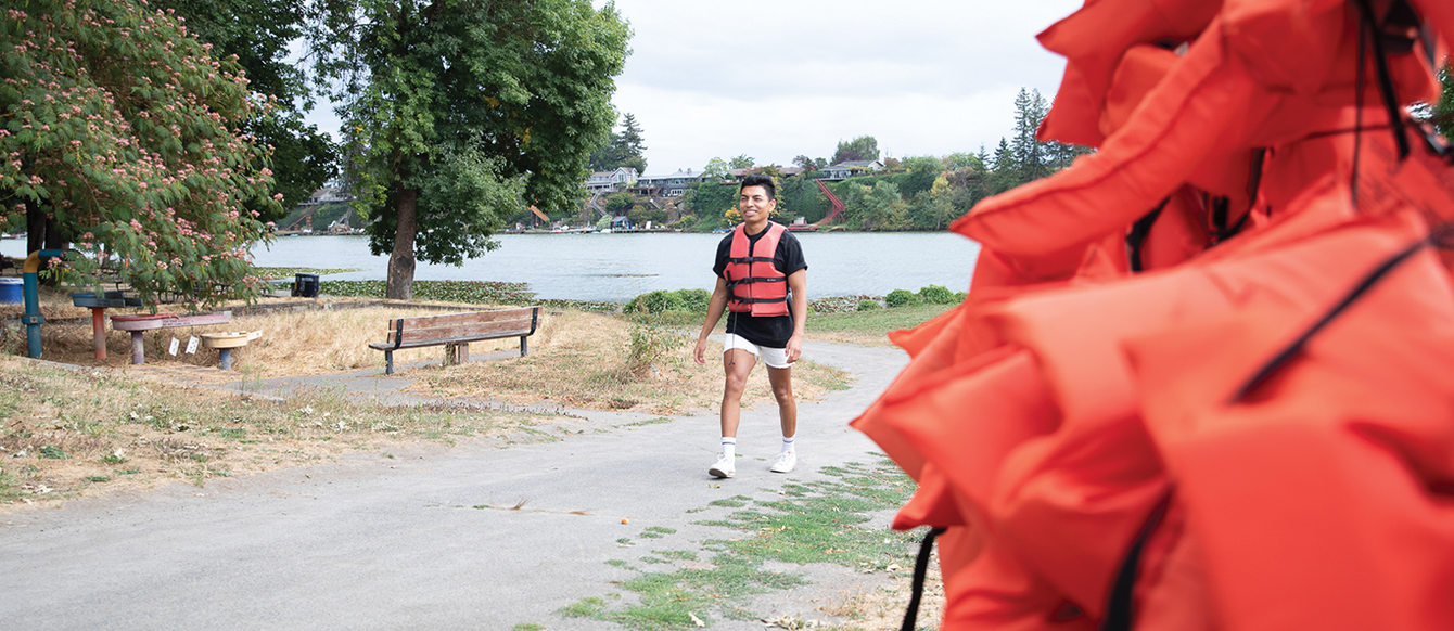 A young man wearing an orange life vest walks up a trail past a collection of life vests. A small lake is behind him.