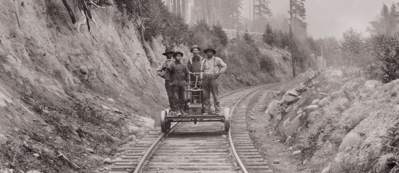 historical, sepia-tone photo of Chinese railroad workers standing on a railroad handcar, one white worker in the middle