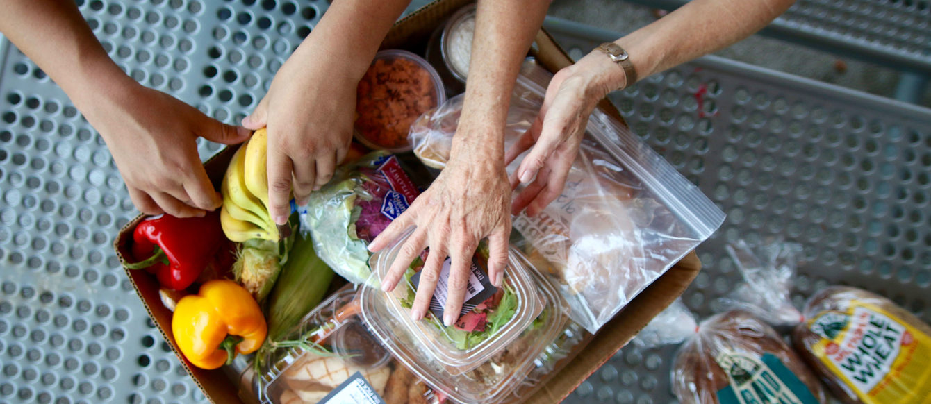 Many hands reach for a box full of food including fresh produce, sandwiches. Two loaves of bread sit on shelf next to the box.