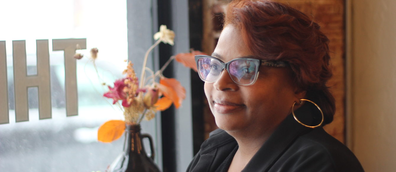 Close up of a woman with cat eye glasses, hoop earrings and a black blazer with auburn hair smiling. A window and vase with flowers are in the background