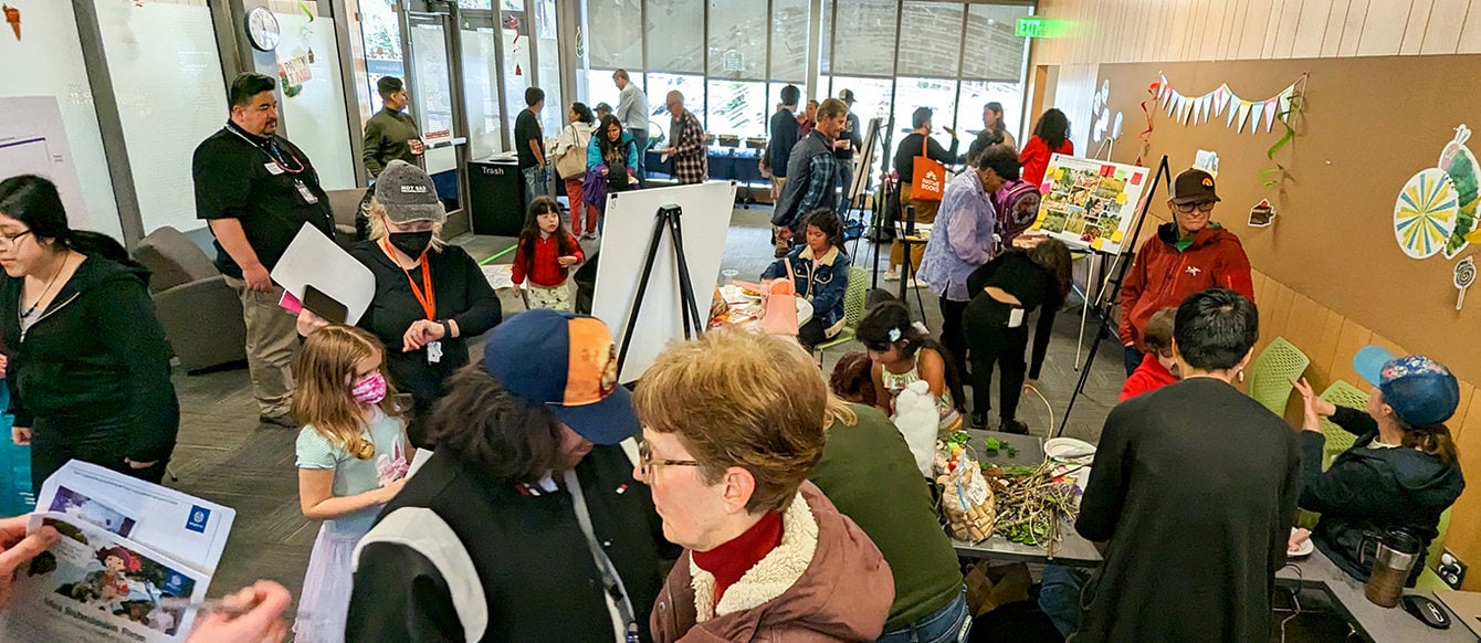 A crowd of several dozen people fill a room with wooden walls and a large bank of windows. The people are engaged in different activities and conversations.