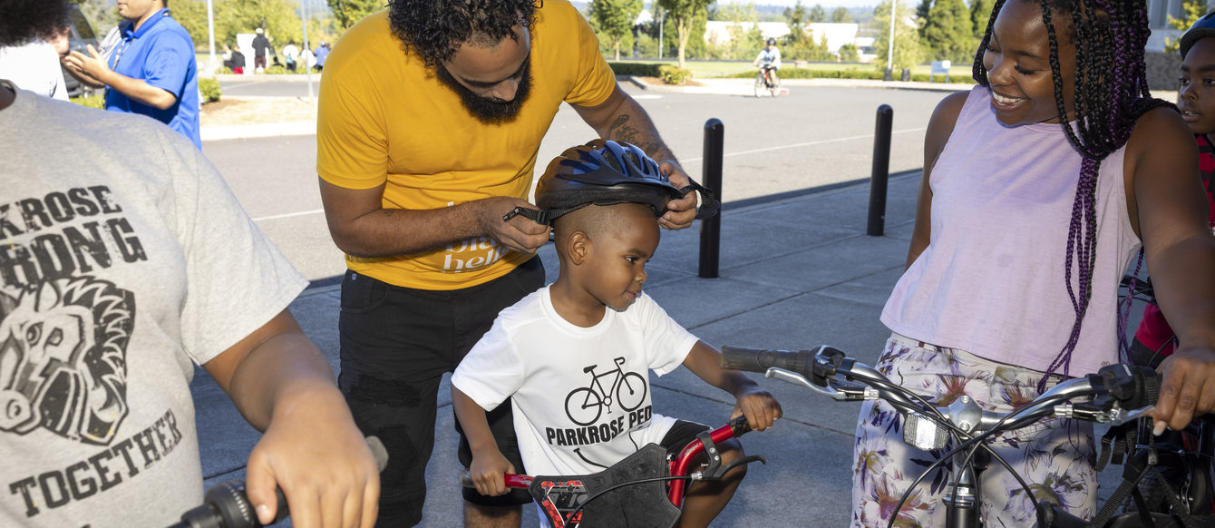 Image of an adult in a yellow t-shirt putting a bike helmet on a child who is sitting on a bike.
