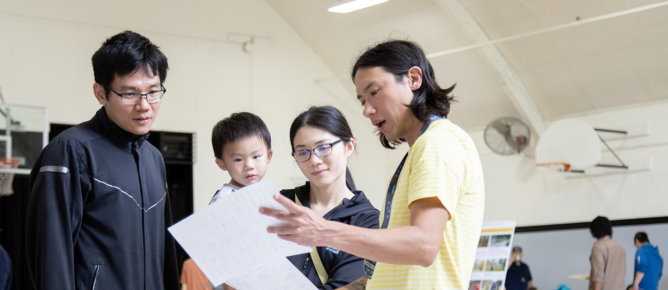 A father, mother and toddler listen to a park designer inside a gym. Other community members are in the background.