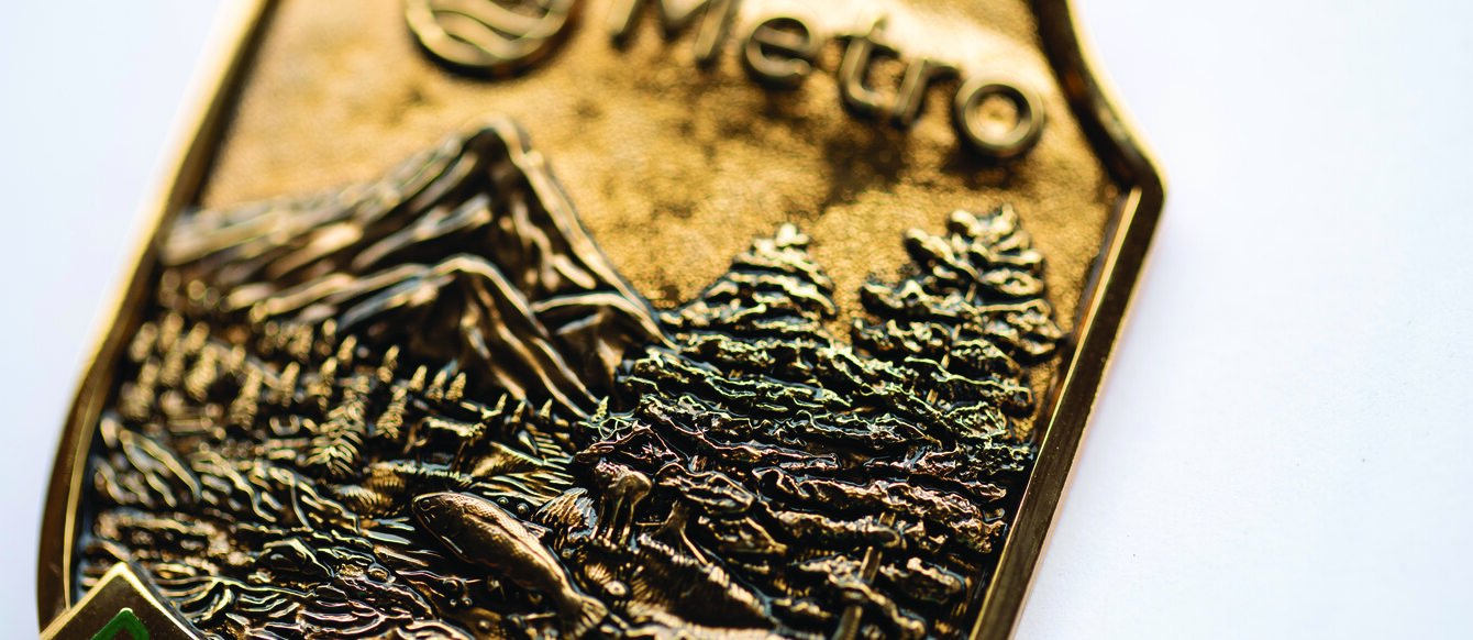 A close up of a bronze park ranger badge has details of mountains, trees and a river. It reads "Metro Park Ranger". 