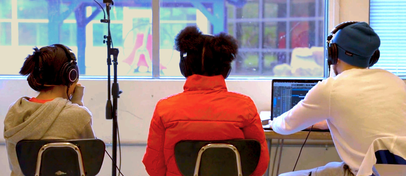 A group of young people sitting in a classroom with headphones on in front of a laptop. A microphone on a stand is nearby.