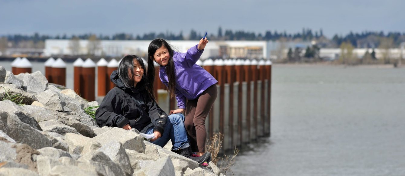 a woman and child sit on rocks in front of a boat ramp's pier on the Columbia River. The child is pointing to something in the distance.