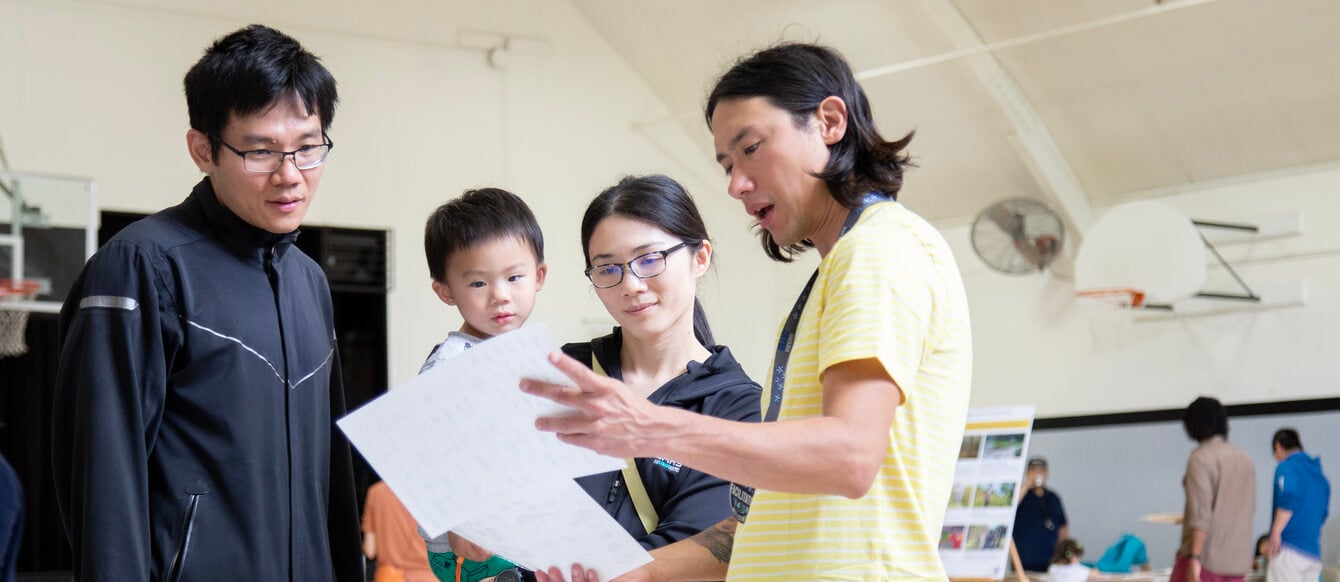 A family, two parents and toddler, are listening intently as an event facilitator speaks to them during a community workshop event.