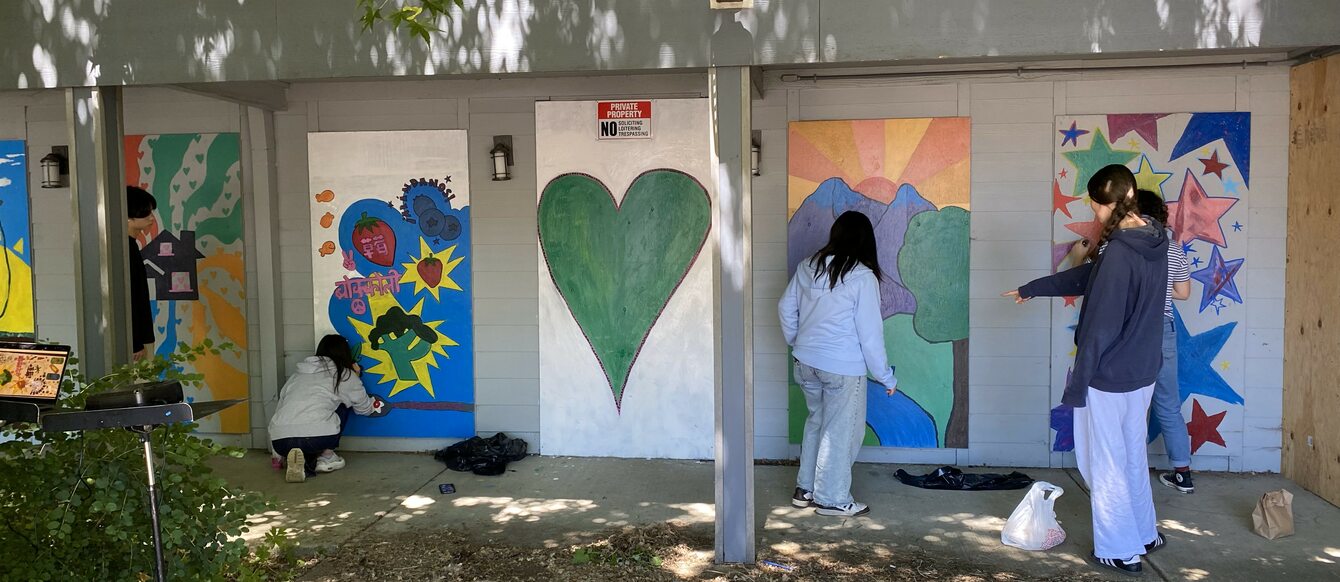 Students work on a mural at the side of a building. The mural includes four panels featuring paintings of vegetables, a large heart, a sunset, and colorful stars.