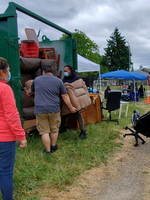 A group of people load pieces of furniture into a large green metal storage unit on a grassy field along a street in Portland
