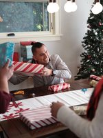 Three people around a dining room table investigating wrapped presents with a Christmas tree in the background.