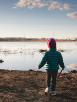 A small child faces away from the camera at the shore of a lake on a sunny winter day.