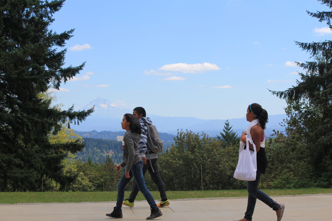 Three young people are walking, shown in profile. They are outdoors surrounded by evergreen trees and grass, with Mt. Hood in the background.