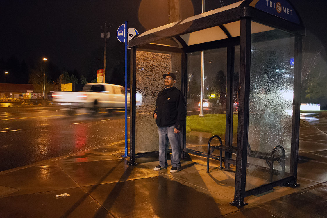 A man waiting for the bus at a bus stop at about 5:30 in the morning.