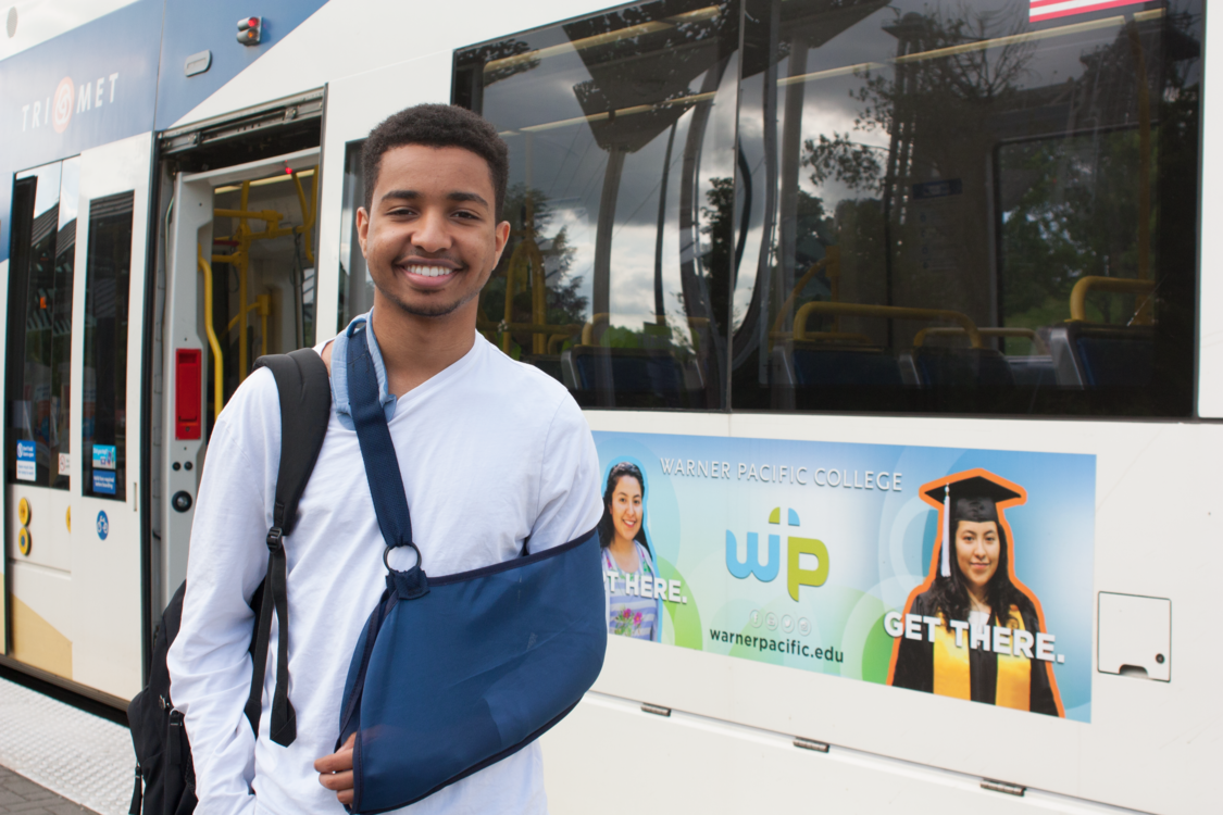 A portrait of 18-year-old Ibrahim Ibrahim standing on the platform of a MAX light rail station with the train in the background.