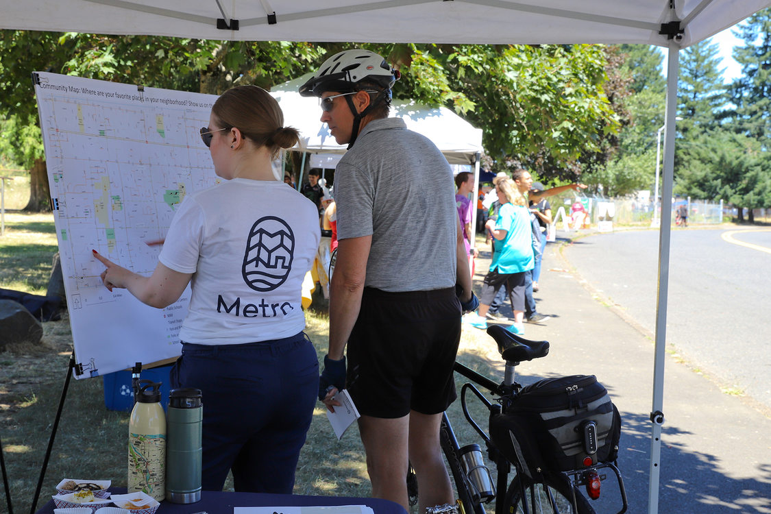 Two people stand in front of a map under a tent at a street fair.