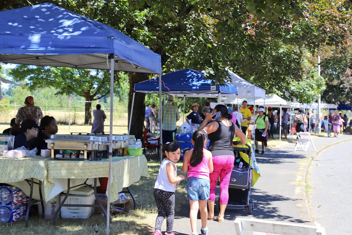 Families walk past park vendors on a tree-lined street.