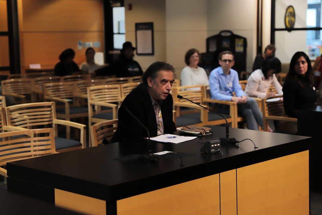 a man speaking into a microphone while sitting a table with a small audience in the background.