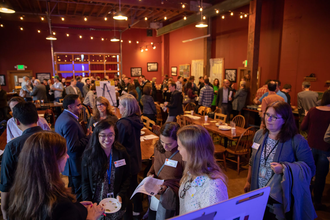 a room full of people standing and talking together in groups; long tables in the center