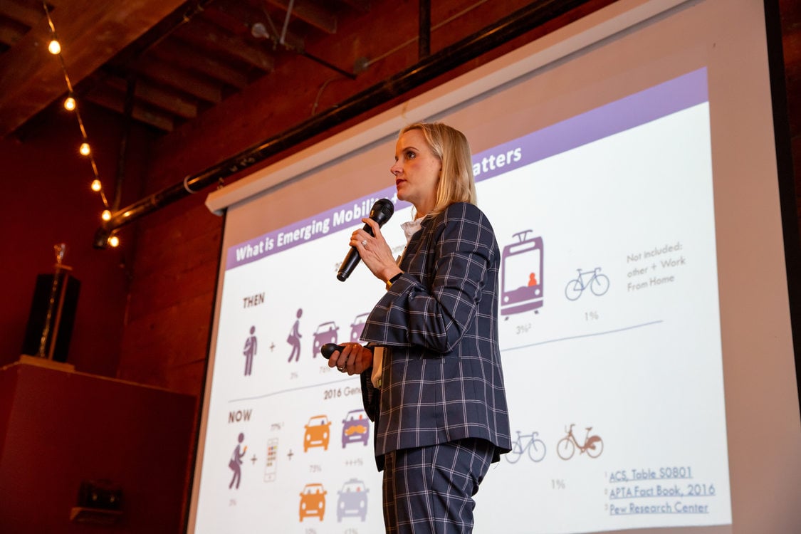 A woman stands in front of a projected screen, delivering a talk as she hold a microphone