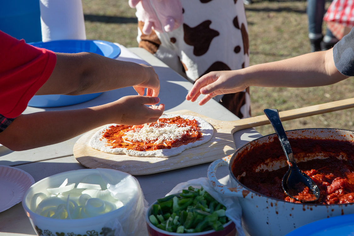 the arms of three little boys adding pizza toppings to a pizza at the center of a table at an outdoor festival