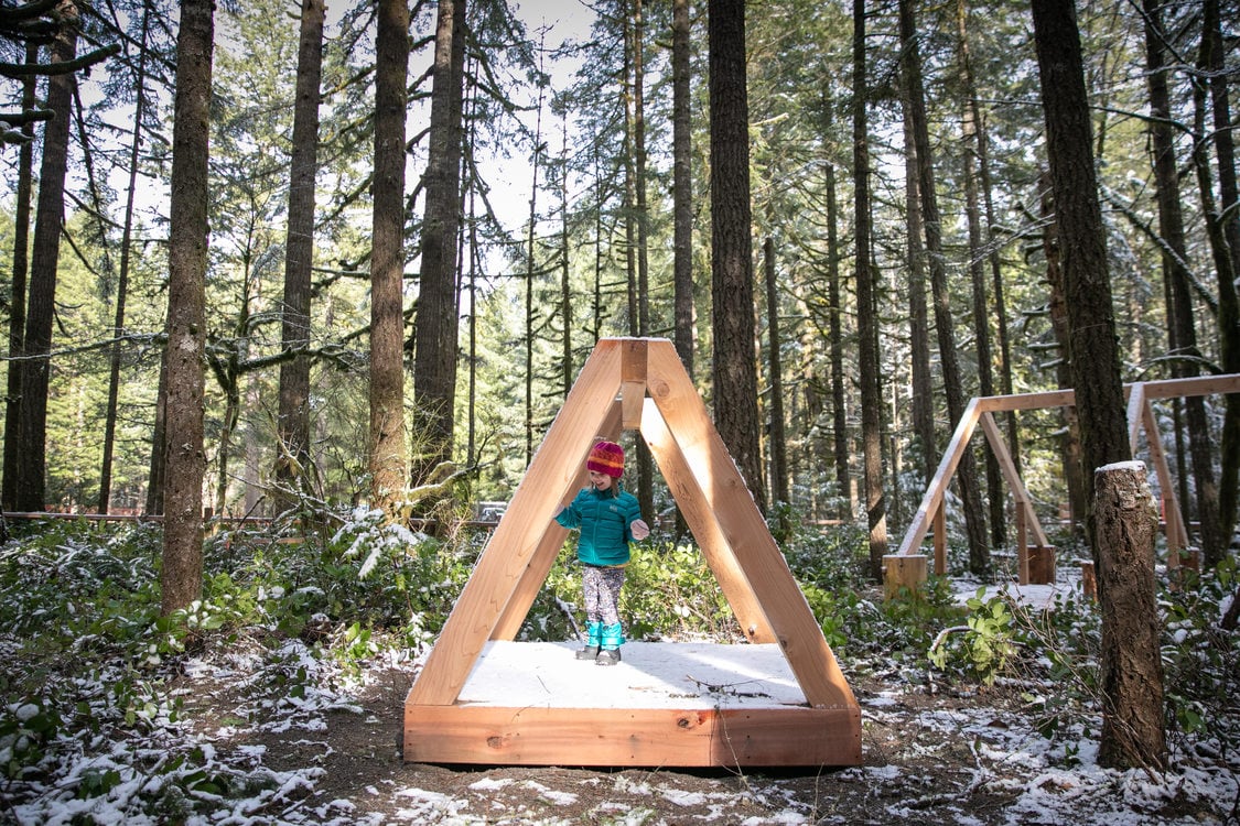 girl in winter clothes stands in triangular nature play structure in woods