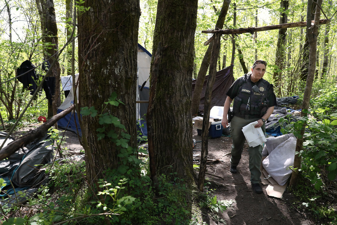 a sheriff deputy, carrying a handful of white garbage bags, visits a campsite in the woods.