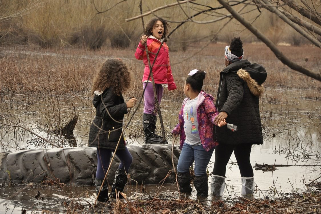 Four youth, ranging in age, stand in a wetland in boot covers. One is facing the rest, standing on a giant submerged tire, holding a large stick. There is marshland visible behind them and tree branches in the foreground.