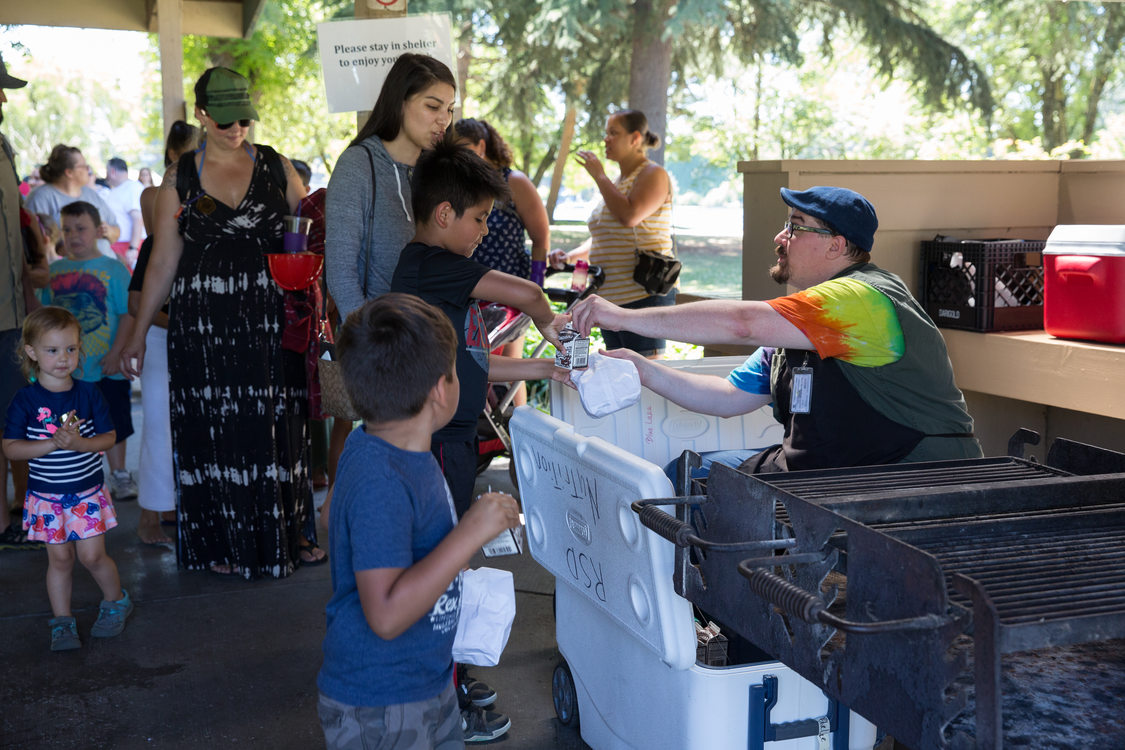 man hands over individual sized milk carton to child in a line receiving free lunch at park