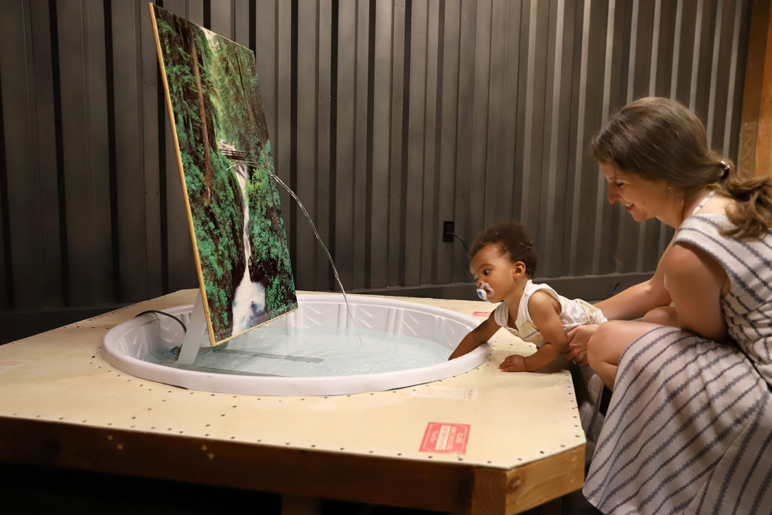a woman holds a baby as it dips his hand into an art installation made out of a plastic wading pool