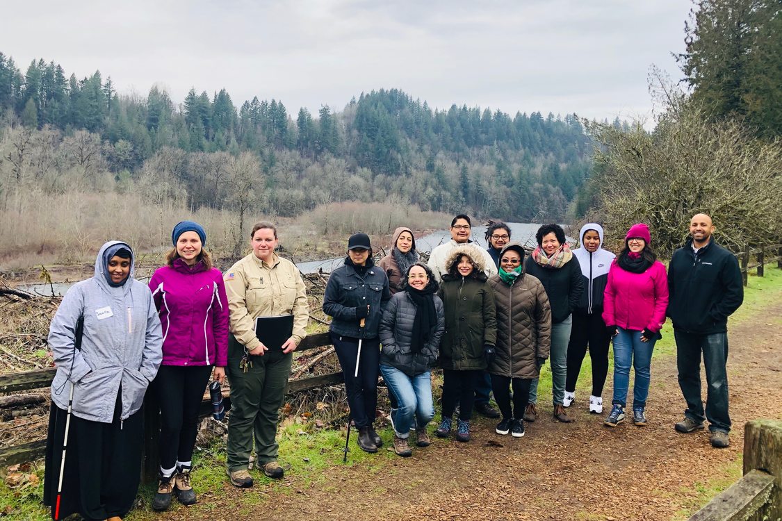 Group of 14 people standing on trail taking group photo with river and forest in background on cloudy winter day