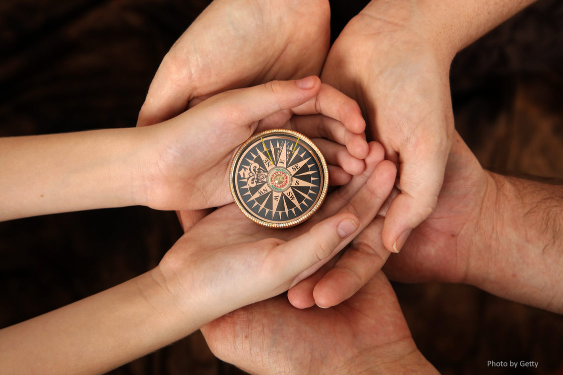 Adult hands cradle the hands of a child.  The child's hands cradle an antique compass. 