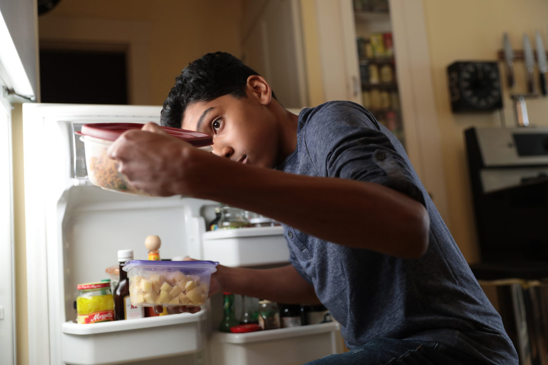A boy opens the refrigerator door and pulls out leftovers stored in reusable containers 