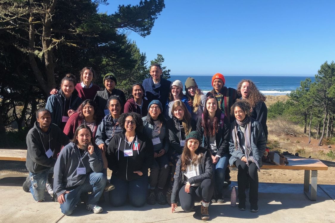 A group of about 15 youth and young adults pose with their instructors in front of some trees with the beach visible in the background.