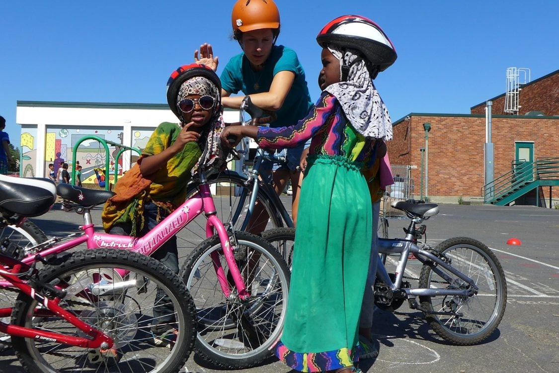 Two girls in head scarves and bike helmets stand and lean on their bicycles on the blacktop of a schoolyard as an adult who is also wearing a helmet is speaking to them.