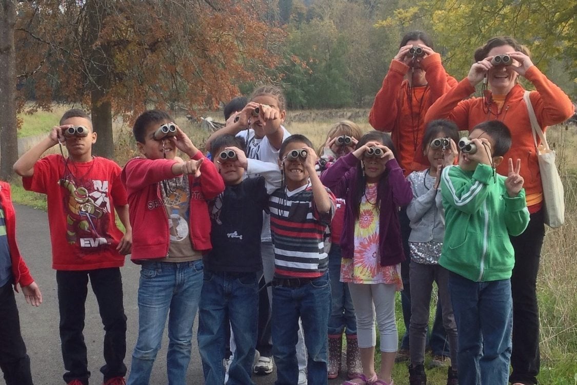 A group of about 8 children and two adults are facing the camera but looking through binoculars at something beyond the photographer with a field and trees in the background.