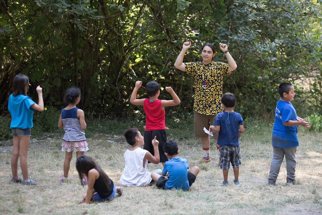 Eight young children with their backs to the camera are in a field with trees in the background focusing on an instructor who is holding their arms up. Two of the children are copying the gesture.