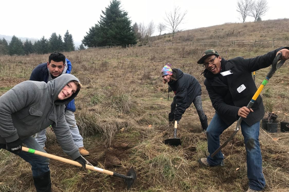 Four teenagers with shovels face the camera and smile while clearing grass in a field with trees visible in the background.