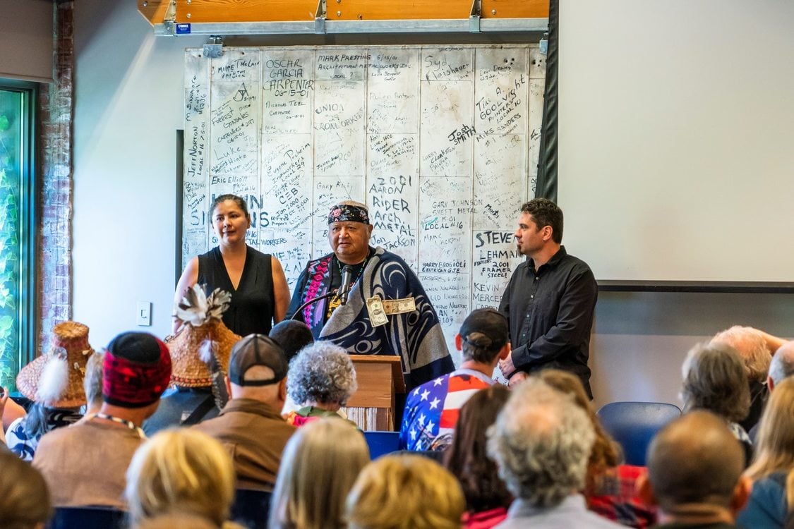 Three adults stand at a podium part of the audience is visible with their backs to the camera.