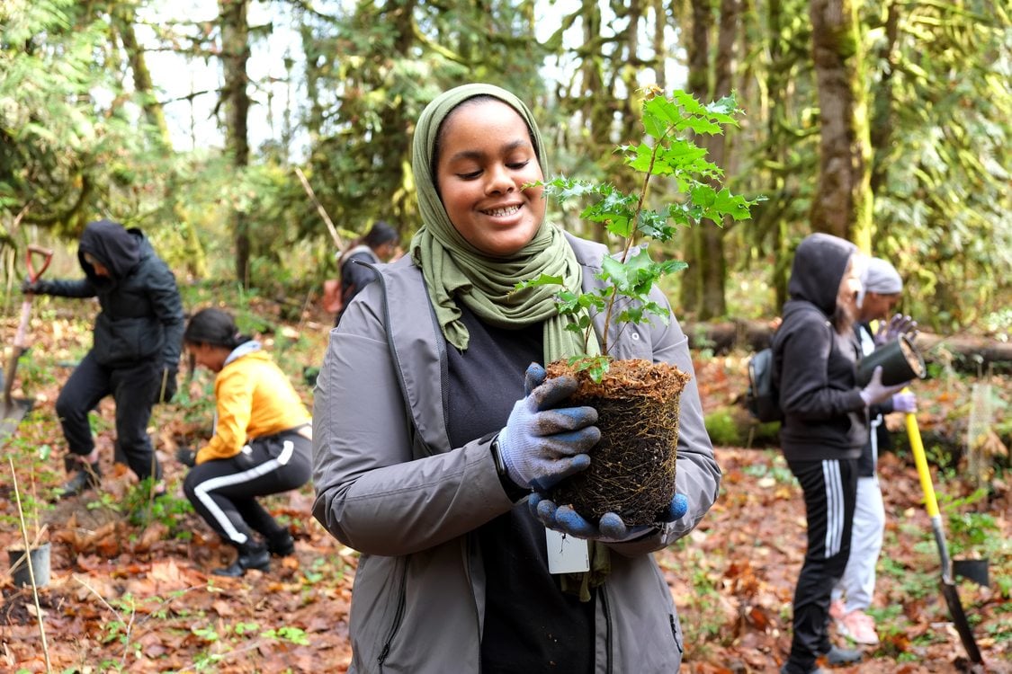 A young woman in hijab holds a tree as other teenagers plant trees at Oxbow Regional Park.