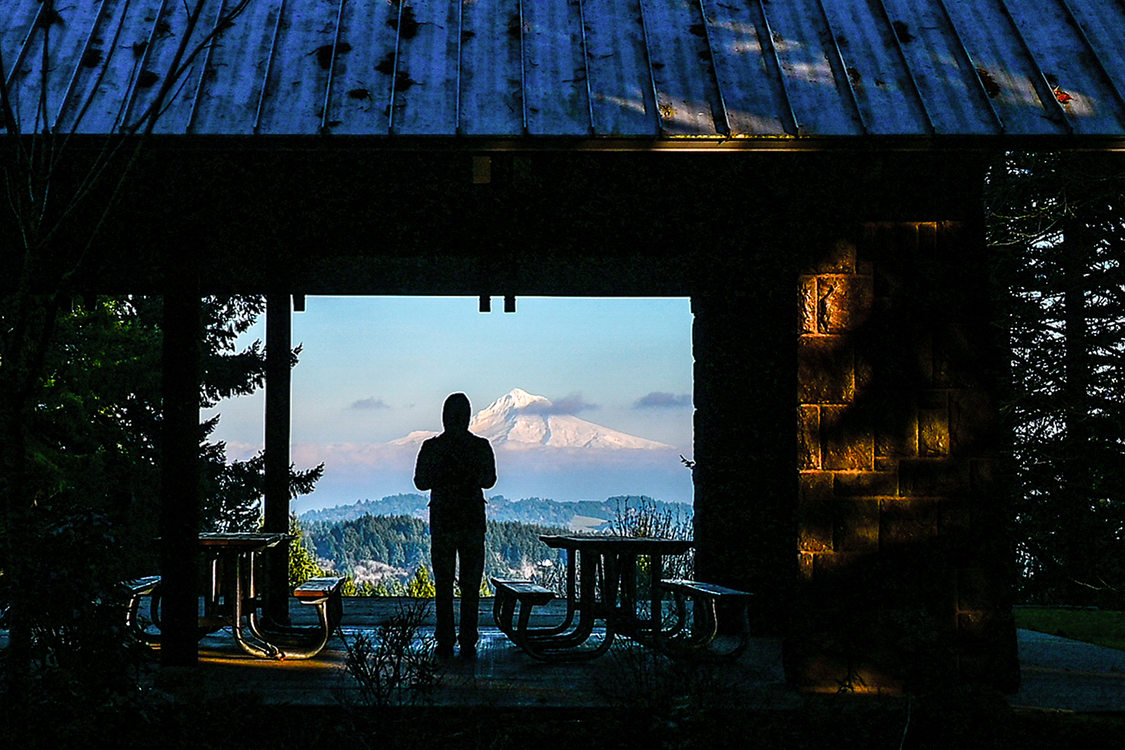 A person stands in the shadow of a picnic shelter while Mount Hood, covered in snow, rises in the horizon.