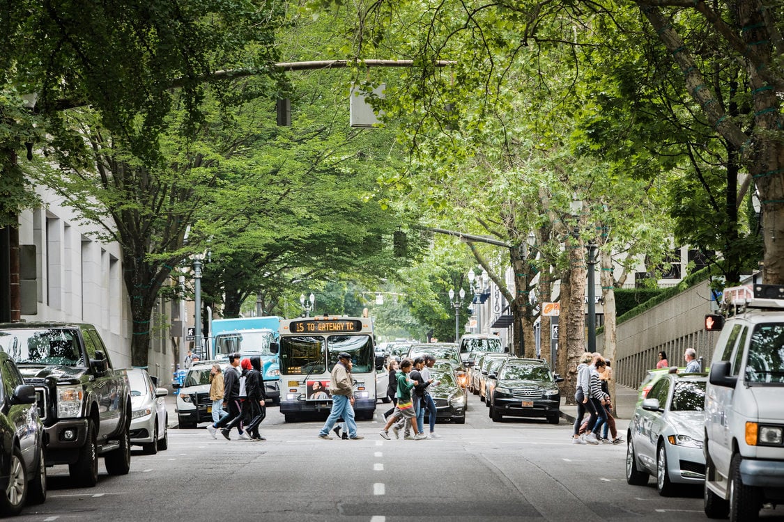 pedestrians cross the street in front of a bus on a street lined with parked cars