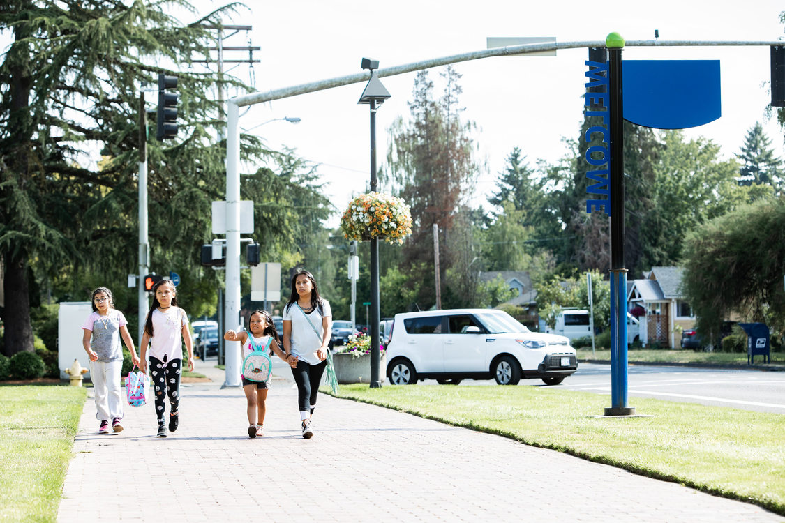 A woman and three children walk on a sidewalk, a car turning in the background