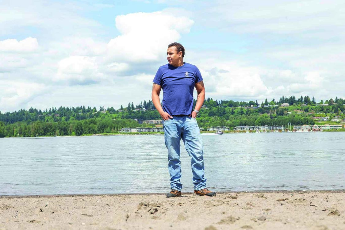 A black man stands on the bank of the Columbia River on a sunny day with blue skies.