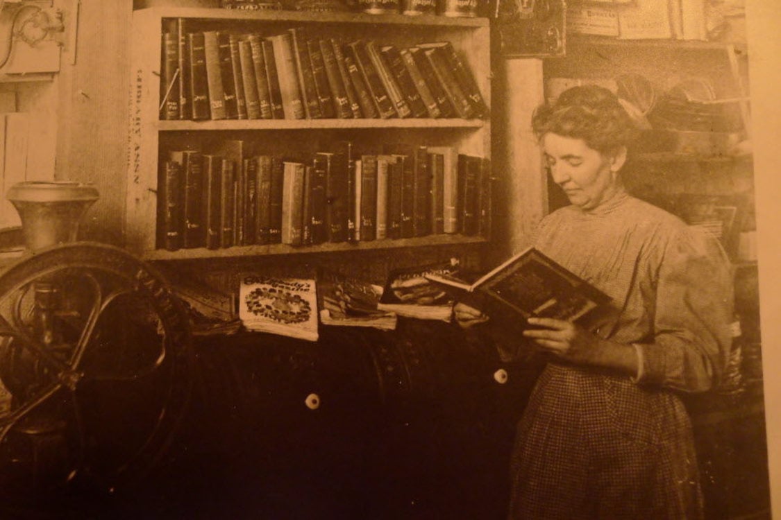A sepia-tone photograph of a woman reading a big, leather-bound book in front of a shelf of other big, leather-bound books.