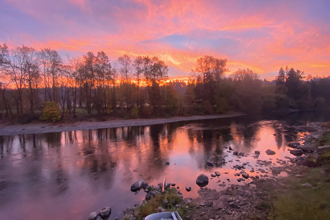 Purples, pinks and oranges fill the sky and are reflected on a river. A small boat sits on the shore.