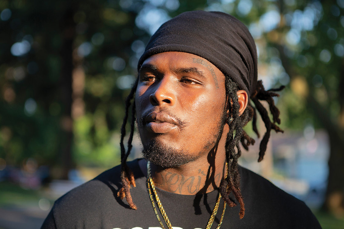 A Black man with twists and a black head wrap poses for a photograph with the setting sun shining on his face.